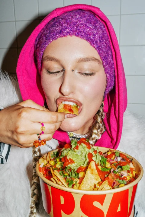 Woman wearing a bright pink hood eating nachos topped with guacamole, salsa and fresh vegetables as part of Salsa Shop’s Live Saucy campaign.