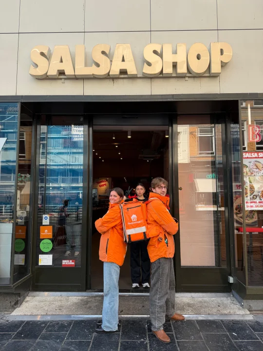 Two Thuisbezorgd delivery riders with orange delivery bags standing outside a Salsa Shop restaurant.